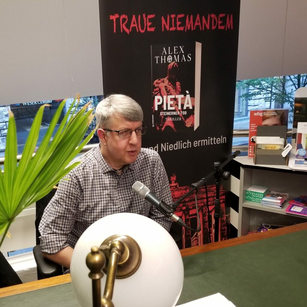 Alex Thomas mit Mikrofon an einem Schreibtisch. Dahinter ein Rollup-Banner mit dem Buchcover von "Pietà - Steinerner Tod" Foto © J. Windolf