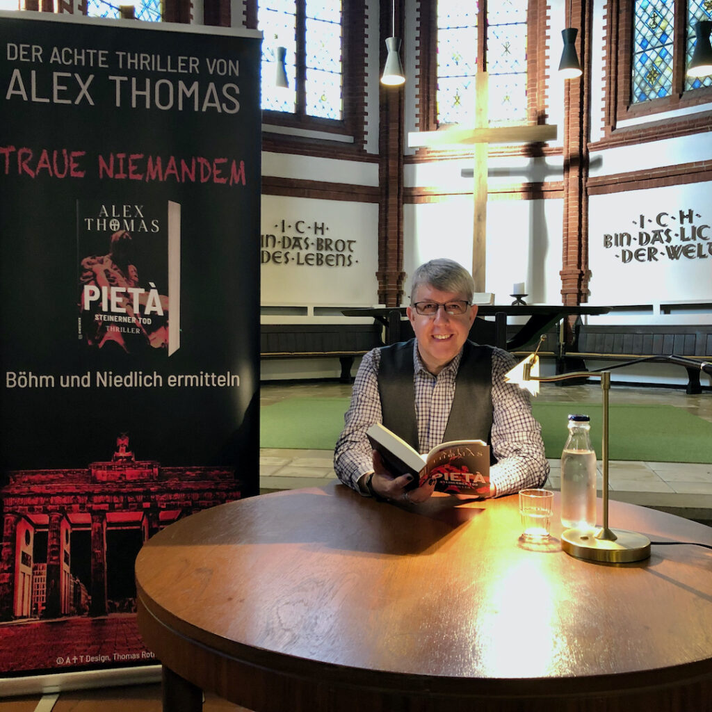 Alex Thomas mit Mikrofon an einem runden Tisch. Dahinter ein Rollup-Banner mit dem Buchcover von "Pietà - Steinerner Tod" Foto © Alex Thomas